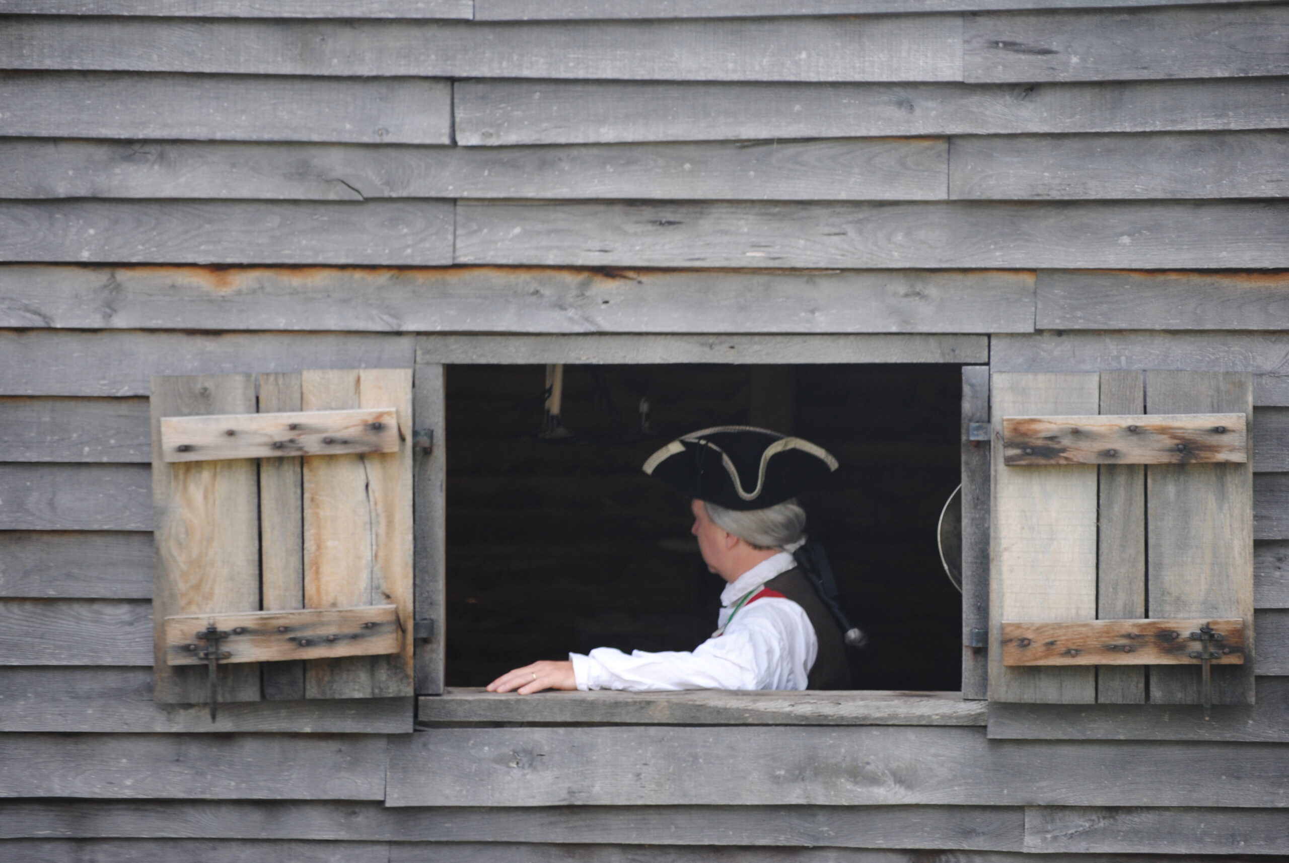 A person in an 18th-century tricorn hat and white shirt leans out of an open wooden window.